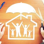 Vue de dessus de mains tenant des ciseaux près d'un découpé en papier représentant une famille sous une maison et un parapluie. L'image illustre l'action de **changer son assurance emprunteur** pour faire des économies, dans le cadre de la Loi Lemoine, tout en conservant une protection familiale équivalente.